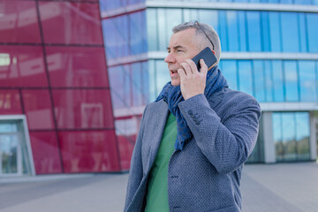 Portrait of concentrated middle-aged man standing near modern building with glass fasade in city, talking on smartphone.