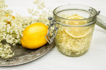 A jar of lemon and fresh elderberry inflorescences in syrup. Vintage metal plate on a white table