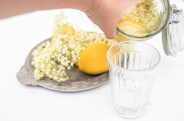 Hand pouring a refreshing summer drink of elderberry and lemon into a faceted glass on a white table.