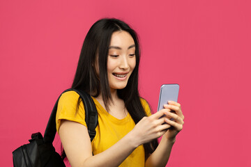 Happy asian female student using smartphone, texting online while standing with backpack on pink background, copy space