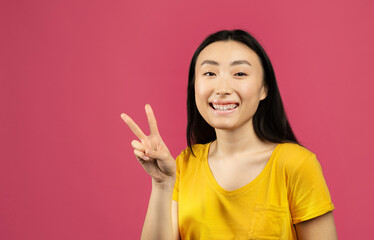 Happy japanese lady gesturing victory symbol and smiling to camera, standing on pink studio background, free space