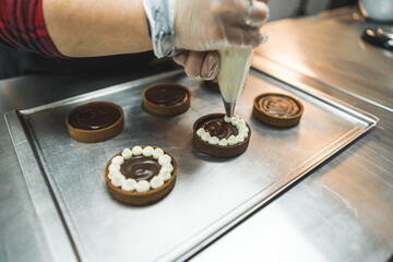Tarts placed on a baking tray being decorated with white cream by a person using a pastry bag. A baker wearing transparent glove. Blurred background. High quality photo