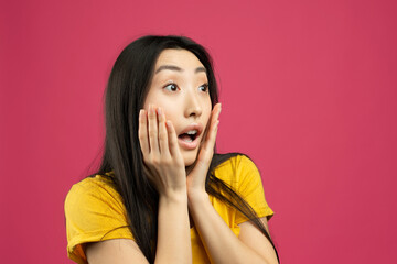 Shocked chinese lady with open mouth holding hands on cheeks, looking at empty space isolated on pink background