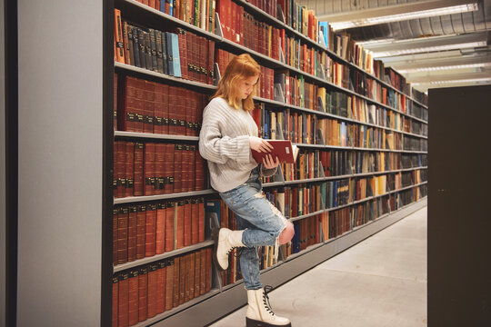 Teen girl with red hair standing by shelves library reading.