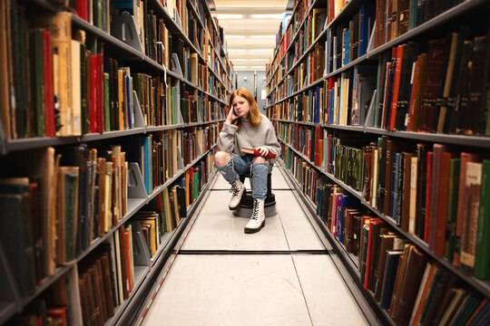 Teen girl sitting with book between library shelves.