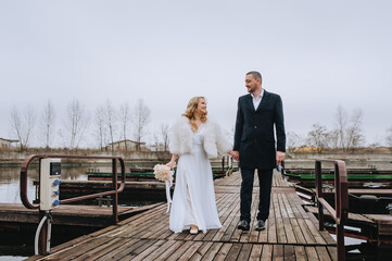 A stylish bearded groom in a black coat and a beautiful curly-haired bride in a white dress, a fur coat with a bouquet of reeds are walking, holding hands, on a wooden pier. wedding photography.