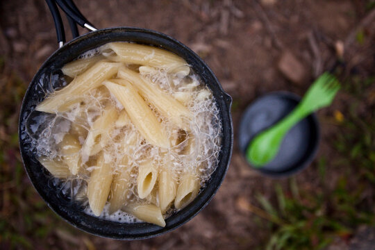 A camping stove boils over with pasta with a camping spoon in the background.