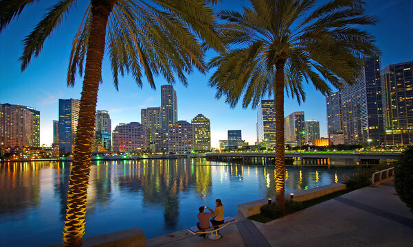 A Couple Takes Picture Of Each Other On A Waterside Park Bench With Views Of Miami At Sunset