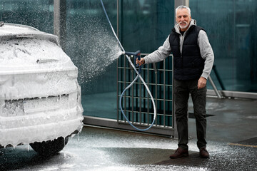 Full-length portrait of Caucasian senior man, smiling looking at camera, washing his car in...