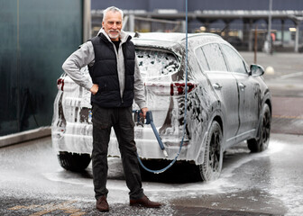 Handsome senior man smiles at camera, poses with high pressure hose in self-service car wash station