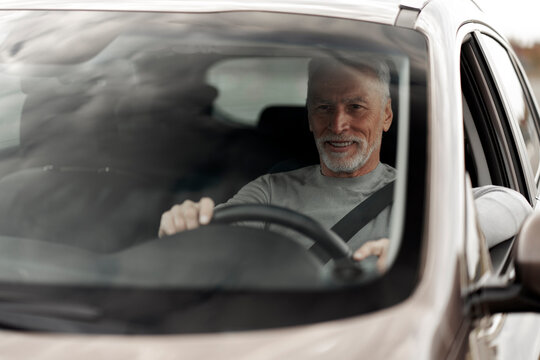 View through windshield to a smiling 60-70 years senior man with fasten safety belt, driving car 