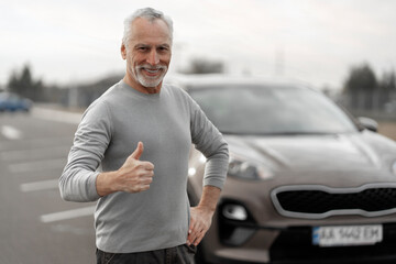 Handsome smiling senior man shows thumb up, standing near his comfortable car on automobile parking