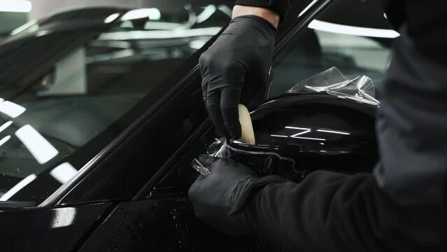 Side view of a mechanic wearing black long sleeve sweatshirt and black gloves applying paint protective film on black car's outside mirror. High quality 4k footage