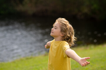 Peaceful kid with raised hands meditating, feeling calm. Kid practice yoga and relaxed on nature. Child faith, praise, happiness and freedom. Kid closed eyes and feeds energy of nature, dreams.