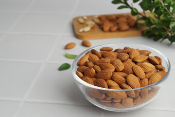Bowl of delicious almonds and fresh leaves on white tiled table. Space for text