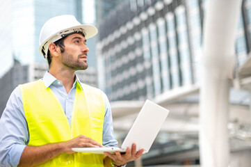 Caucasian man use laptop to working  and wearing a yellow vest and hard hat at the construction site in the city.