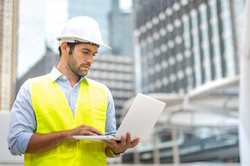 Caucasian man use laptop to working  and wearing a yellow vest and hard hat at the construction site in the city.