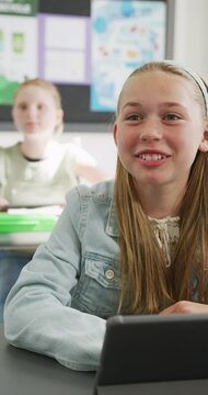 Vertical video of happy caucasian schoolgirl sitting at desk, talking in class, copy space
