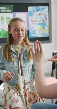 Vertical video of happy caucasian schoolgirl using sign language with teacher in class, copy space