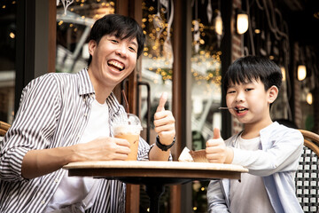 Happy Asian father and son look at camera with free time cheerful at cafe. Self portrait of young father and his little son smiling while sitting on the chair. Lifestyle and happiness.