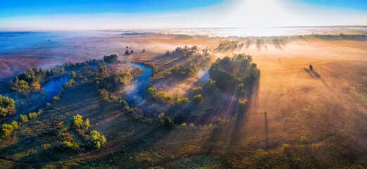 The channel and bends of the river, on a marshy meadow. Orange dry grass, scorched by the summer heat, and morning fog. A wonderful landscape at dawn. Drone view.