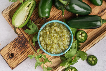 Wooden board with bowl of tasty green salsa sauce, jalapeno peppers and cilantro on light background