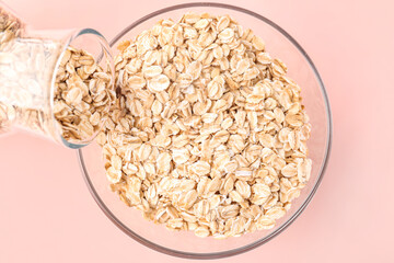 Pouring of raw oatmeal from bottle into bowl on pink background, closeup