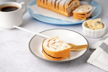 White plate with piece of delicious sponge cake roll, cup of coffee and chocolate on white table