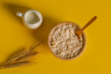 Bowl with oatmeal, spoon, wheat ears and pitcher on yellow background