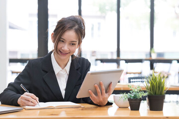 Beautiful Asian business woman using her laptop to work and enjoy working, taking note, reviewing assignment and smiling in the office.