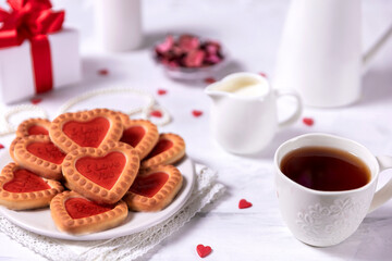 Fototapeta na wymiar Homemade cookies in the form of hearts with red jam on Valentine's Day. On a marble table with a gift, tea and white candles.