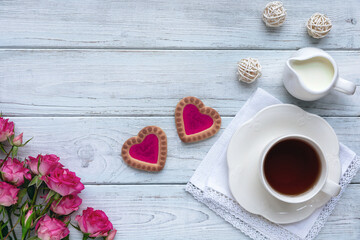 Homemade cookies in the form of hearts with pink jam with a cup of tea on wooden light blue boards with roses. The concept of Valentine's Day. Flat lay.