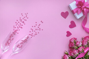Two champagne glasses with confetti, roses and a gift box on a pink background. Flat lay, top view, copy space.