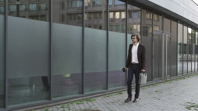 A businessman with a briefcase walks near a large office building and looks around. Young manager, technology, leader. The concept of success.