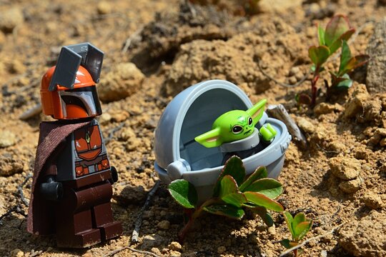 LEGO Mandalorian with small Yoda alien race Child called Grogu in flying pod standing next to green young plants on arid desert like soil. 