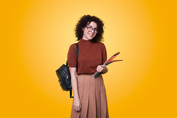 Young curly student woman wearing backpack and eyeglasses holding books isolated over yellow background. School education. Modern style.