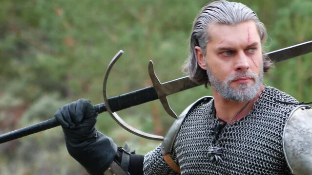 Portrait of a medieval warrior with gray hair and a scar on his face in chain mail with shoulder pads and a two-handed sword in his hands posing against the backdrop of the forest. Witcher Middle Ages