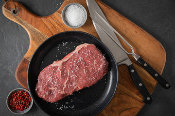 Entrecote Steak roh vom Rind mit Meersalz und roter Pfeffer auf Teller bei Vorbereitung in Küche mit Messer, Fleischgabel und Holz Brett auf Schiefer Stein Hintergrund Schwarz