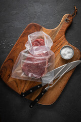 Entrecote Steak roh vom Rind in Plastik Vakuumbeutel für Vakuum marinieren und Sous Vide garen mit Meersalz auf Holzbrett in Küche mit Messer, Fleischgabel und Schiefer Stein Hintergrund Schwarz