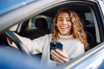 Beautiful young woman is sitting inside a car on the drivers seat with phone. Business, taxi,...