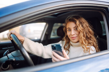 Naklejka premium Beautiful young woman is sitting inside a car on the drivers seat with phone. Business, taxi, technologie, internet, online concert.