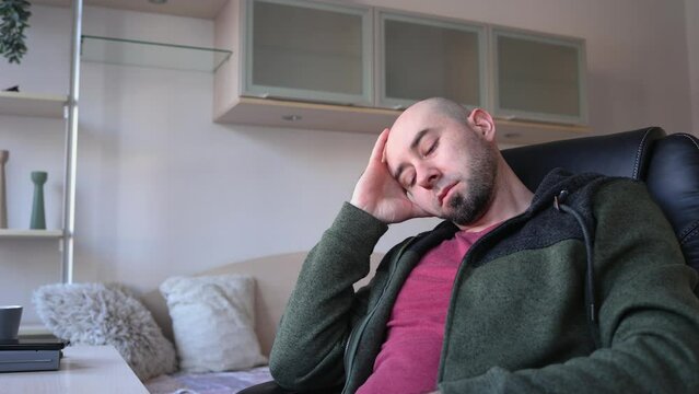 Young Caucasian bearded bald man sitting on chear and thinking looking at window. Depressed person in living room. Psychology and mental health.