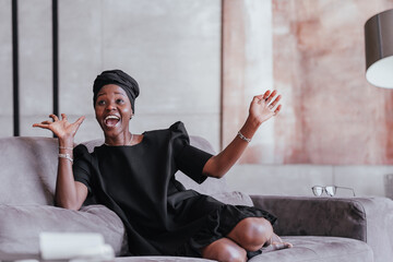 Excited African young adult woman in black dress and turban shouts out loud sitting on cozy couch...