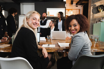 Two attractive young colleagues with different ethnicity sitting at desk looking at camera smiling. Team working in workplace.