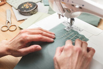 Fototapeta premium Tailoring Process - Women's hands with her sewing. Close up view of sewing process. Female hands stitching fabric on manufacturing machine at workplace.