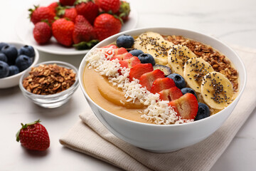 Delicious smoothie bowl with fresh berries, banana, coconut flakes and granola on white table,...