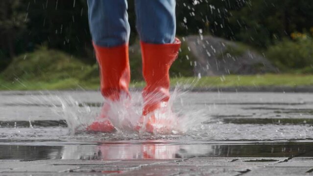 Woman in bright orange rain boots jumping into puddle outdoors, closeup