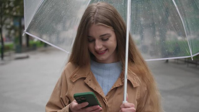 Young beautiful woman with umbrella using mobile phone while waiting someone outdoors
