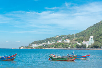 Fototapeta na wymiar Wooden fishing boats with Vietnamese flags anchored near shoreline at Bai Truoc beach with mountain cable cars and hotel buildings in background, Vung Tau, Vietnam