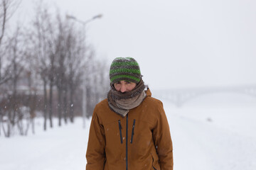 Man in knitted hat and scarf in fitness jacket staying in the snow background in the park in winter